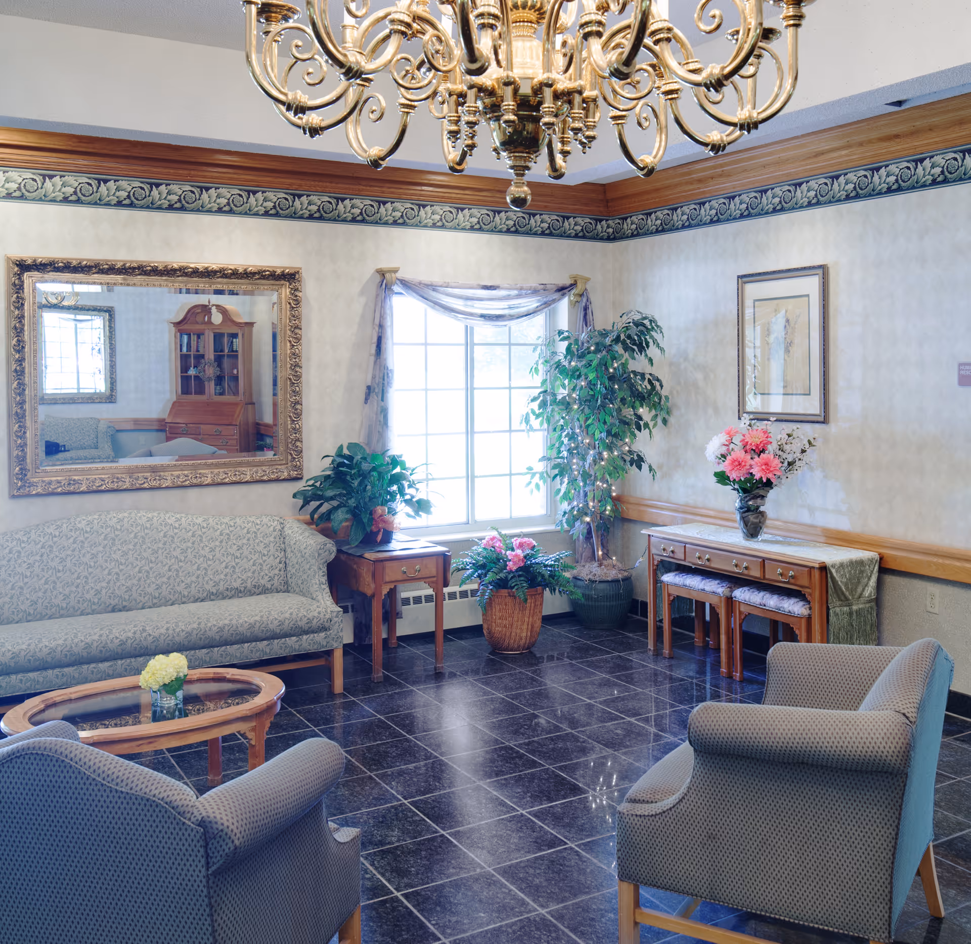 A well-lit living room area with a patterned sofa, two armchairs, a wooden coffee table with a small flower arrangement, and a side table with a plant. There is a large window with sheer curtains, several potted plants, a decorative mirror on the wall, a framed picture, and a wooden console table with a vase of flowers underneath a chandelier.