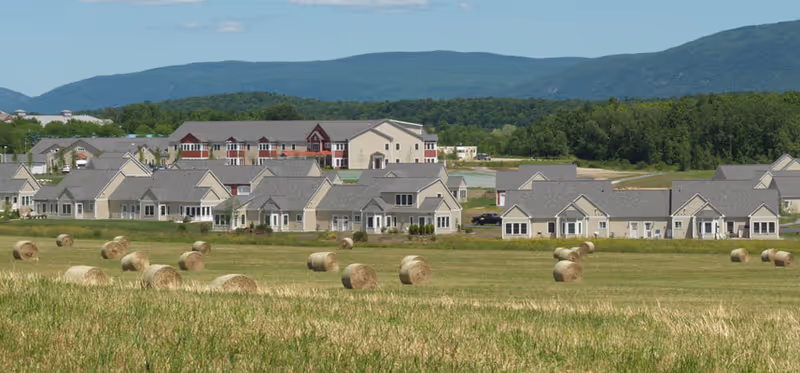 View of a senior living facility named Eastview at Middlebury with multiple single-story and multi-story buildings surrounded by green fields with hay bales, set against a backdrop of forested hills and blue sky.