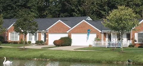 Brick single-story residential units with attached garages and a white picket fence beside a pond with a swan.