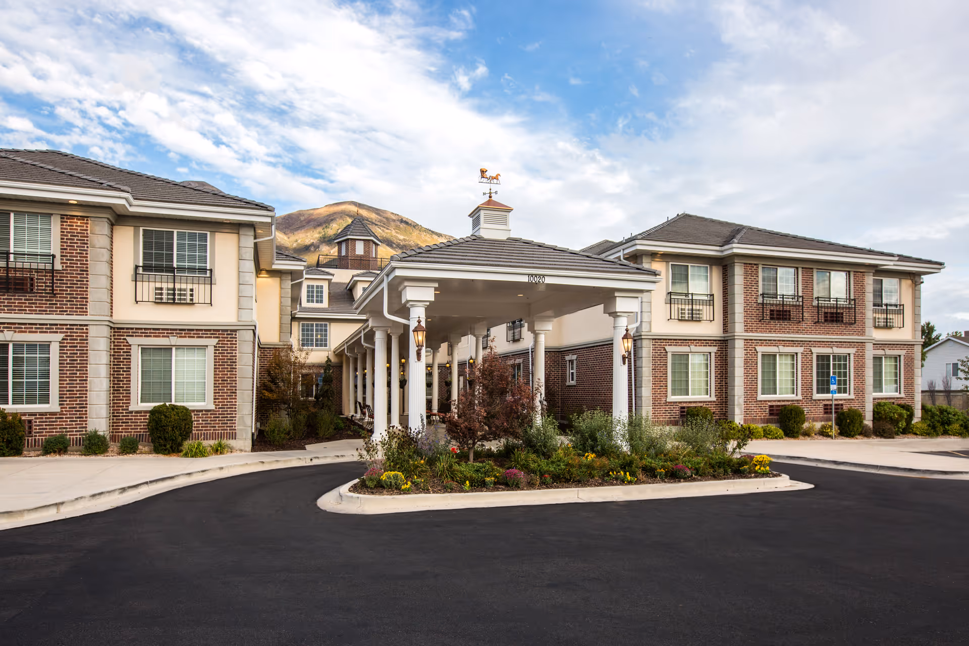 Front exterior view of a senior living facility named The Charleston at Cedar Hills, featuring a covered entrance with white columns, brick and beige siding, landscaped flower beds, and a mountain in the background under a partly cloudy sky.