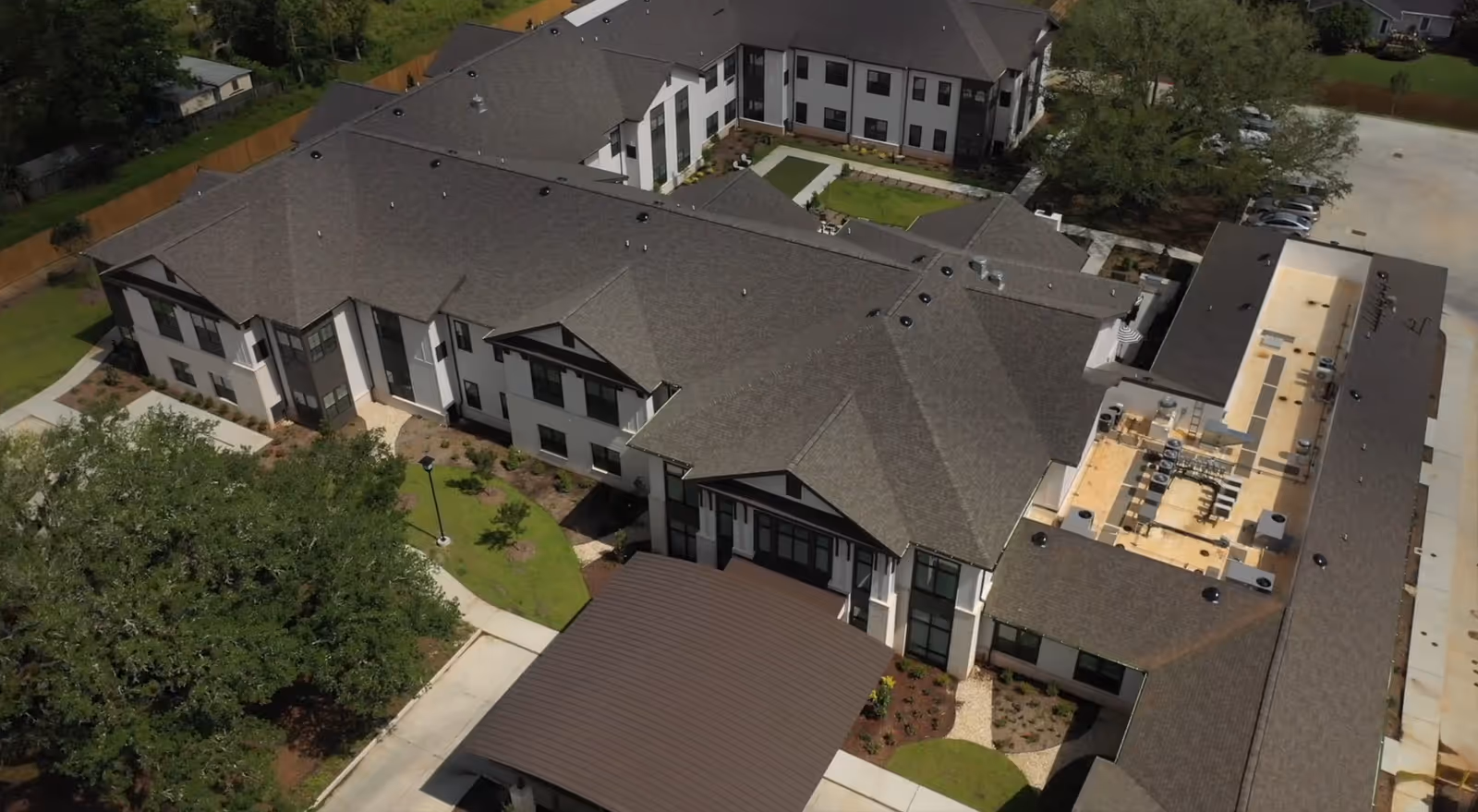 Aerial view of a large senior living facility building with multiple wings, surrounded by landscaped greenery and paved walkways. The building has a dark shingled roof and white exterior walls with black window frames. There is a covered entrance visible at the front.
