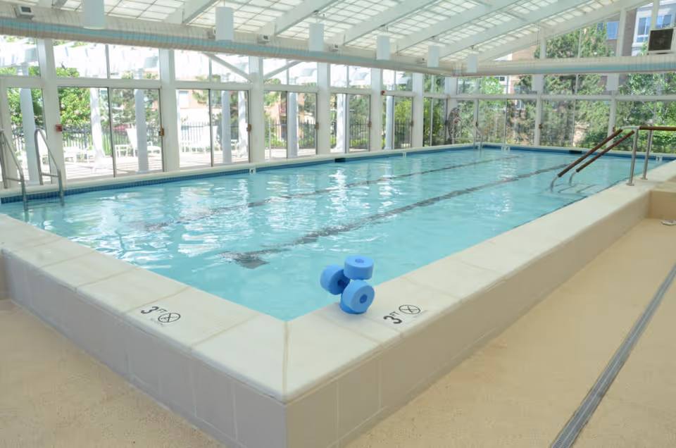 Indoor swimming pool with clear blue water, surrounded by large windows letting in natural light. The pool has depth markings of 3 feet and a pair of blue foam dumbbells resting on the edge. There are metal handrails for entering the pool and a spacious beige tiled floor around it.