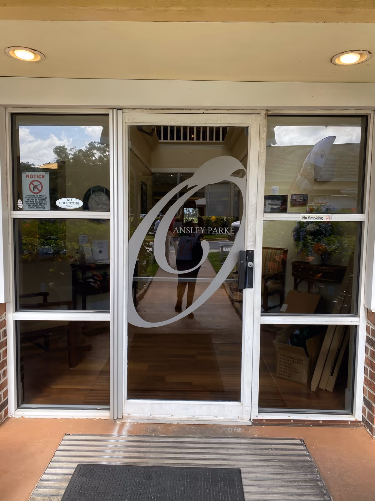 Glass entrance door to Ansley Parke at Oakbridge facility with large decorative letter 'A' and the facility name on the door. The door is flanked by two glass panels with signs including 'No Soliciting' and 'No Smoking'. The interior visible through the glass shows chairs, a table with flowers, and some boxes.