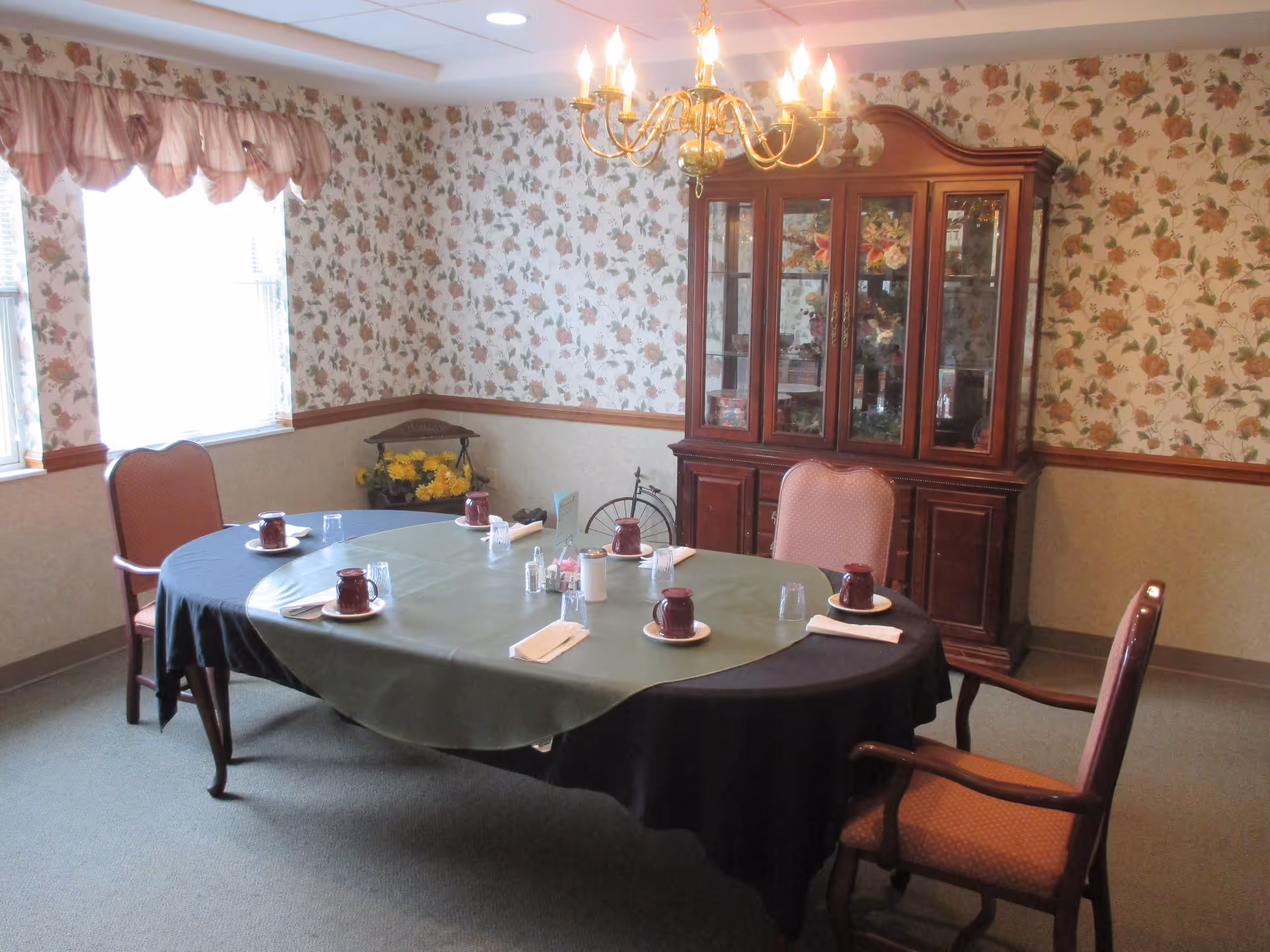 A dining room with a round table covered by a green and black tablecloth, set with four place settings including maroon cups, saucers, napkins, and glasses. The room has floral wallpaper, a wooden china cabinet with glass doors displaying decorative items, a chandelier hanging from the ceiling, and two windows with pink valances letting in natural light.