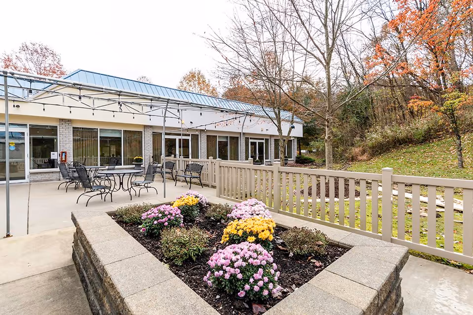 Outdoor patio area at The Haven at North Hills Senior Residence featuring metal tables and chairs, a raised flower bed with pink and yellow flowers, a beige railing, and a backdrop of trees with autumn foliage.