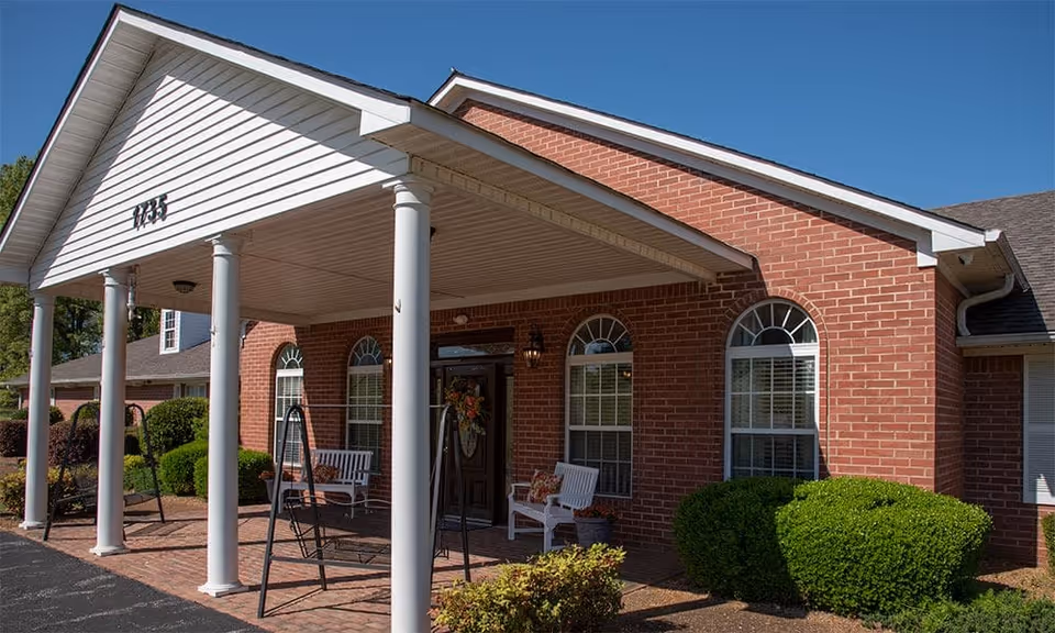 Exterior view of a brick building with a covered entrance supported by white columns. There are two white benches with cushions and two black metal swings on the brick patio. The building has arched windows with white trim and a clear blue sky in the background.