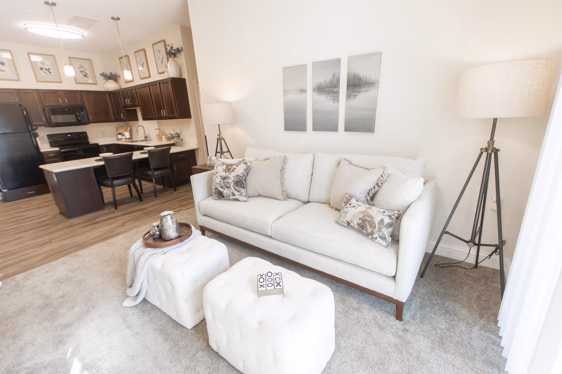 Bright and modern living room area with a light-colored sofa adorned with decorative pillows, two white tufted ottomans, a floor lamp with a tripod base, and a kitchen with dark wood cabinets and black appliances in the background.