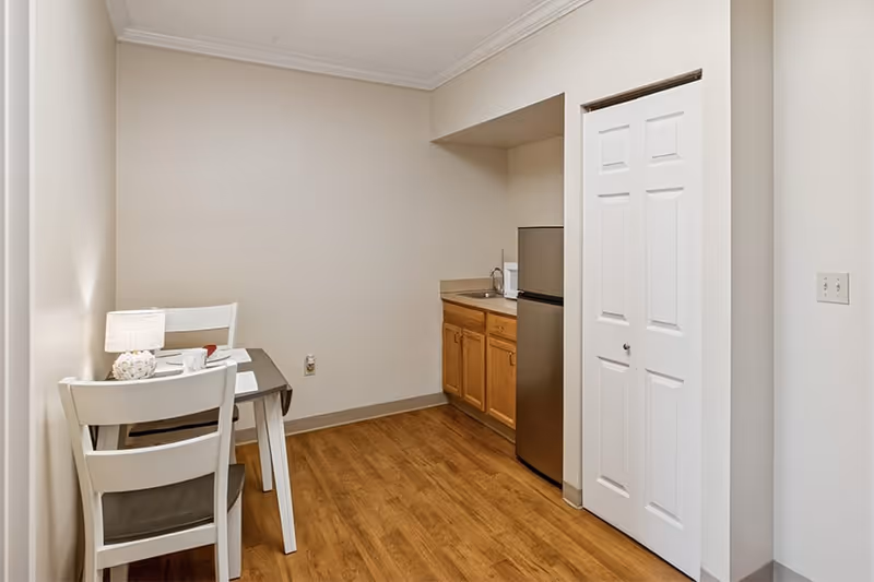 Small kitchenette area with wooden cabinets, a stainless steel mini refrigerator, and a sink. To the left, there is a small dining table with two white chairs and a table lamp. The floor is wood, and the walls are painted beige.