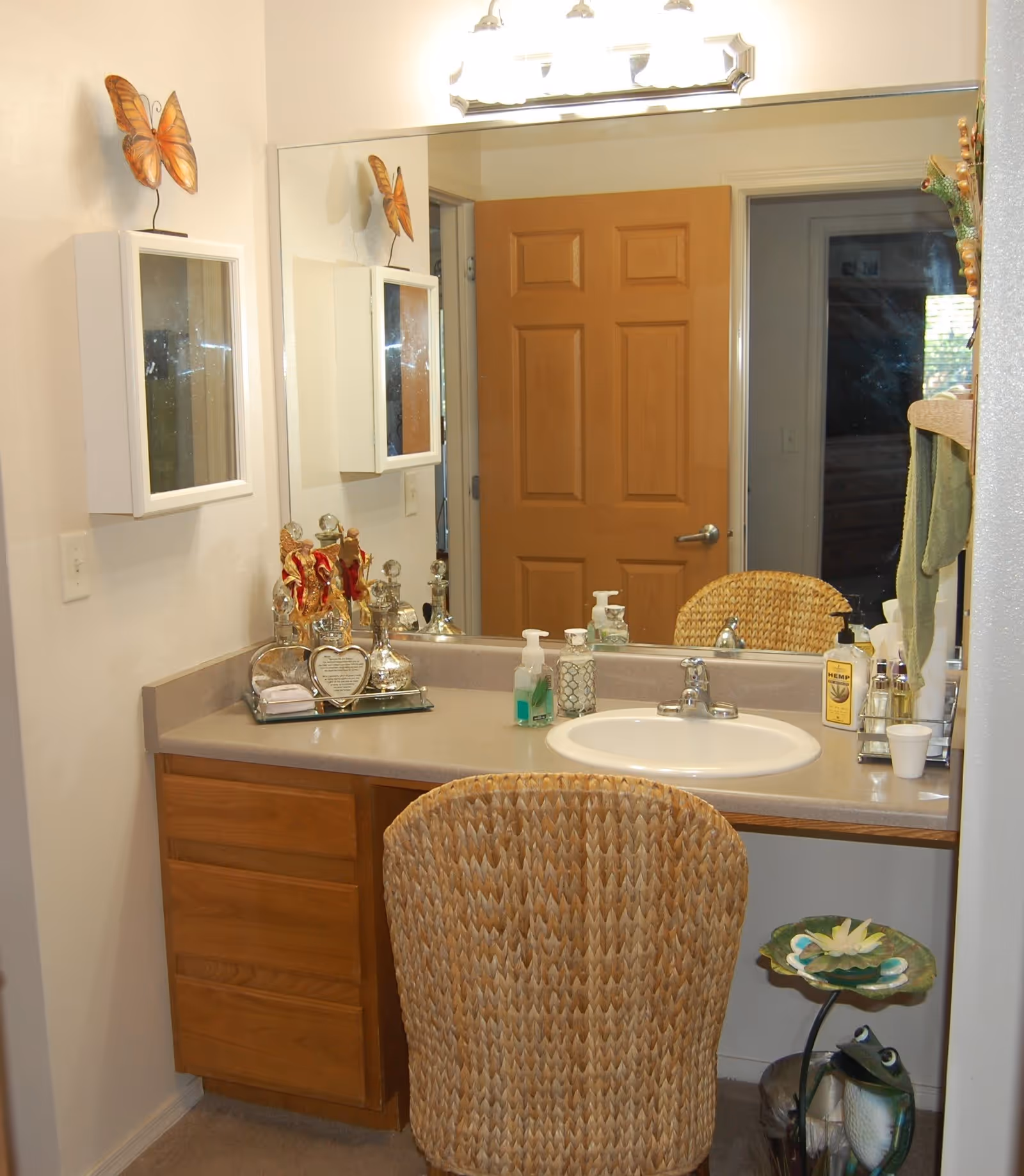 A bathroom vanity area with a large mirror above a countertop that has a sink, soap dispensers, and decorative items. There is a wicker chair in front of the vanity, a wooden door reflected in the mirror, and a small decorative frog and flower stand on the floor to the right.