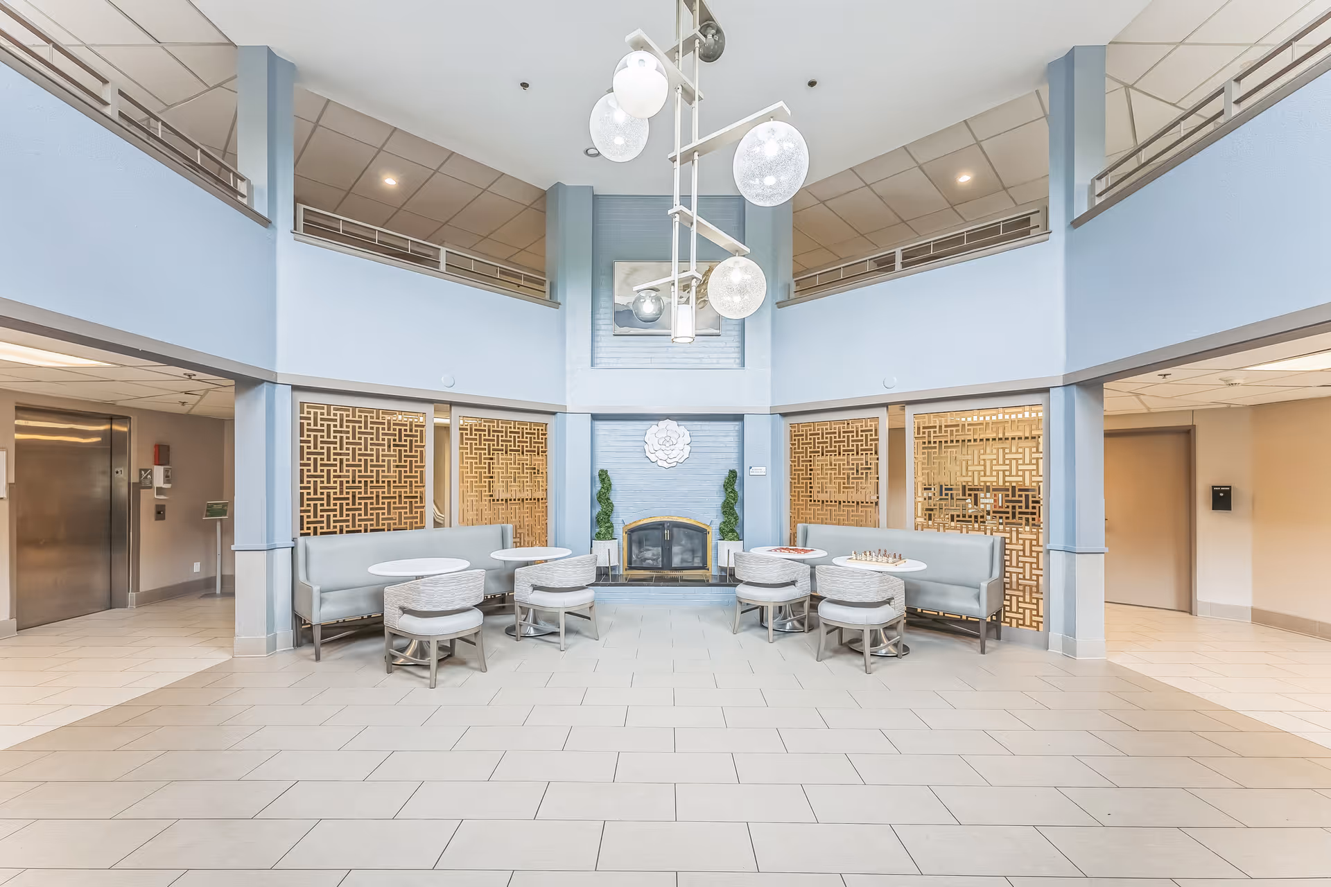 A spacious, well-lit common area with light blue walls and a high ceiling. The room features a fireplace in the center with decorative plants on either side and a framed artwork above it. There are two seating areas with light gray cushioned benches and round white tables, each accompanied by matching cushioned stools. Modern spherical pendant lights hang from the ceiling. To the left, there is an elevator and a hallway, and to the right, another hallway with a door and a mailbox on the wall.