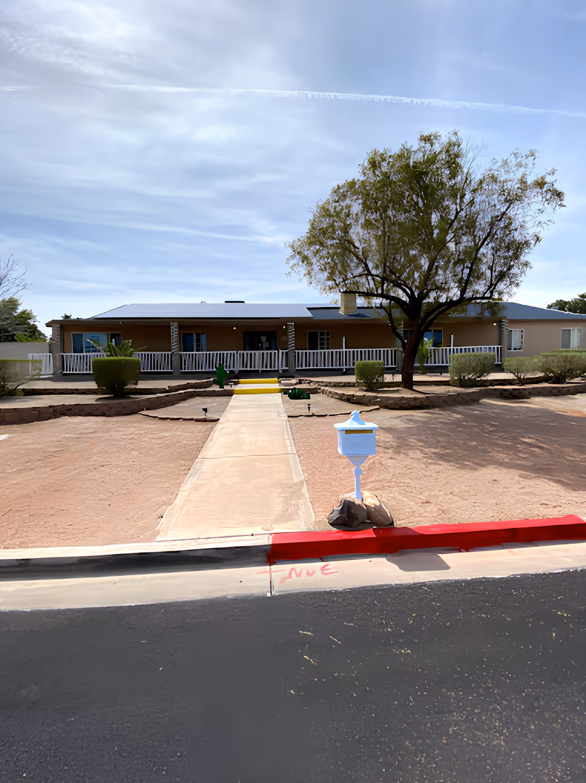 Front exterior view of a single-story building with a wide porch and white railing. A concrete walkway leads from the street to the porch steps, flanked by desert landscaping with trimmed bushes and a tree. A white mailbox is positioned near the curb, and the sky is partly cloudy.