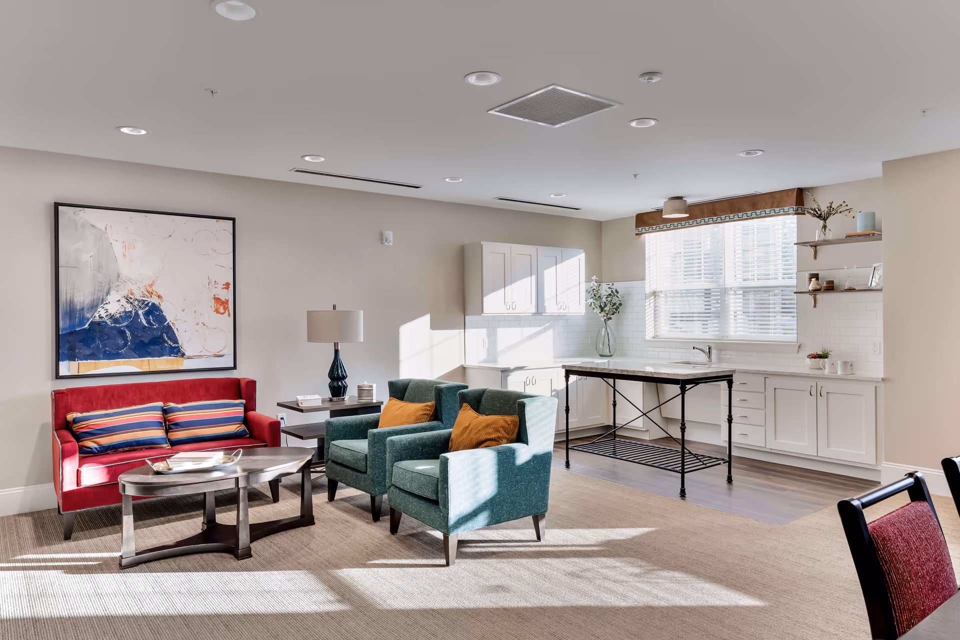 A bright and spacious living area with a red loveseat, two teal armchairs with orange pillows, a dark wooden coffee table, and a side table with a lamp. The background features a white kitchen with cabinets, a window with blinds, and a small island table. The room has beige walls and carpeted flooring.