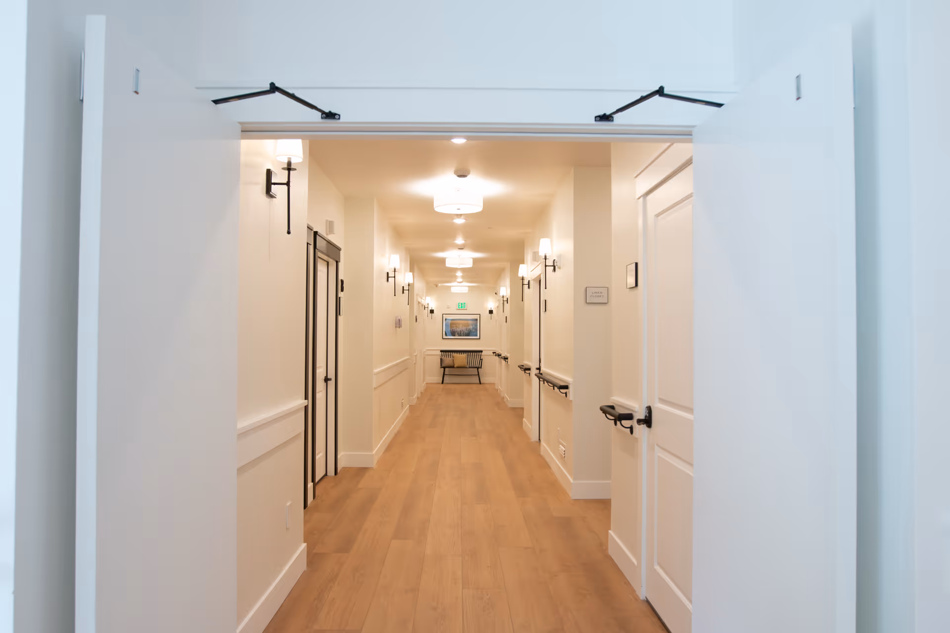 A well-lit hallway in a senior living facility with wooden flooring, white walls, multiple closed doors on both sides, wall-mounted handrails, and several wall sconces and ceiling lights. At the end of the hallway, there is a bench beneath a framed picture and an exit sign above a door.