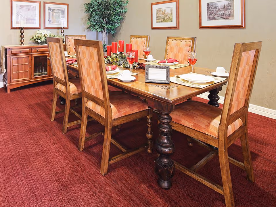 A dining room with a wooden table set for six people. The table has six wooden chairs with cushioned seats and backs in a patterned fabric. The table is decorated with red candles, a centerpiece with greenery and berries, and place settings including plates, napkins, cups, and glasses with red liquid. The room has red carpet flooring, beige walls, framed artwork on the walls, a wooden sideboard with candles and a flower arrangement, and a tall green plant in the corner.