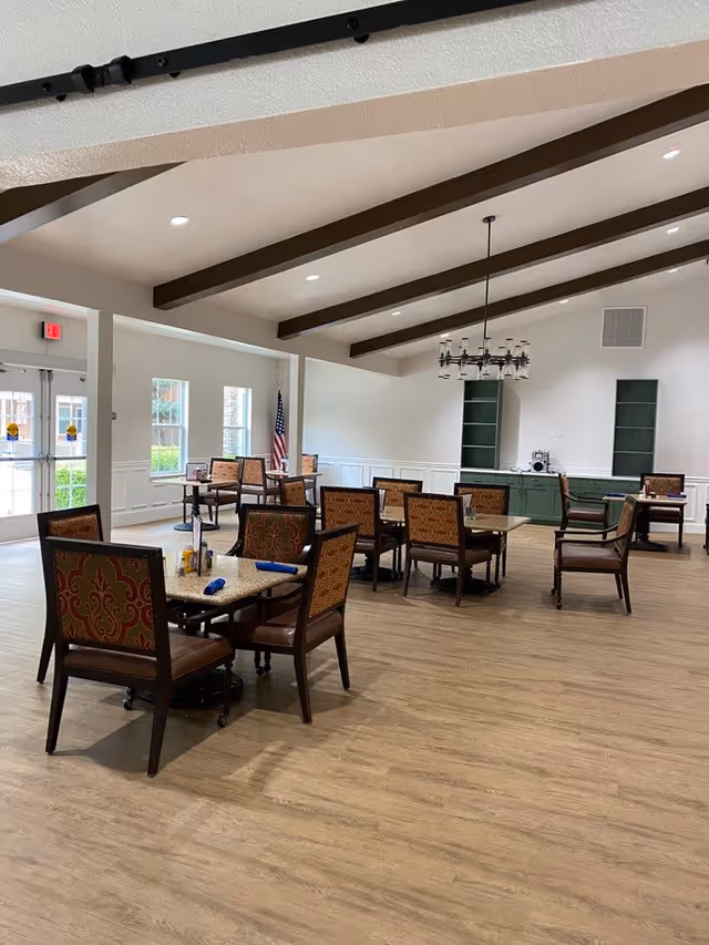 A spacious dining room with several tables and chairs arranged neatly. The room features wooden flooring, white walls with wainscoting, and exposed wooden ceiling beams. There is a chandelier hanging from the ceiling, a green cabinet with shelves against the far wall, and an American flag near the windows. Large windows and glass doors allow natural light to enter the room.