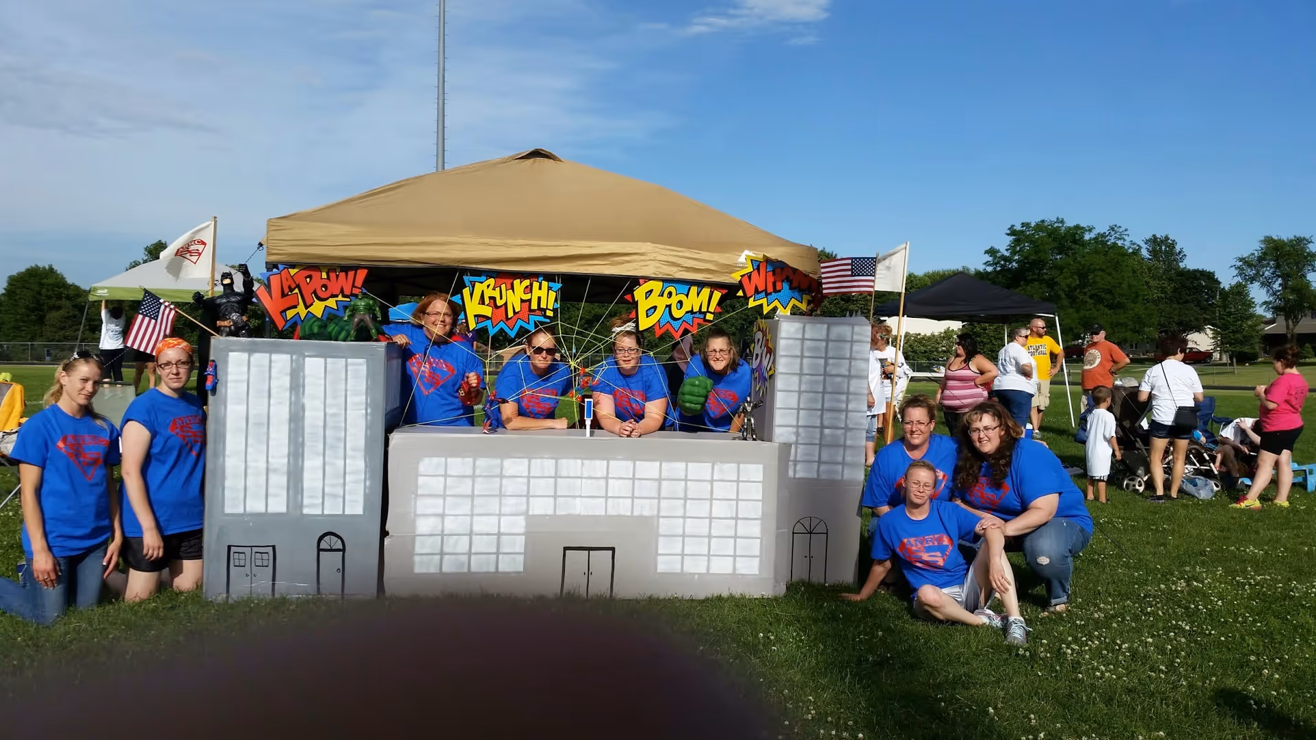 A group of people wearing matching blue t-shirts with a red logo are gathered around and behind a cardboard display designed to look like a city skyline with comic book style sound effects like 'KAPOW!', 'KRUNCH!', 'BLOW!', and 'WHAM!'. They are outdoors on a grassy field with other people and tents visible in the background under a clear blue sky.