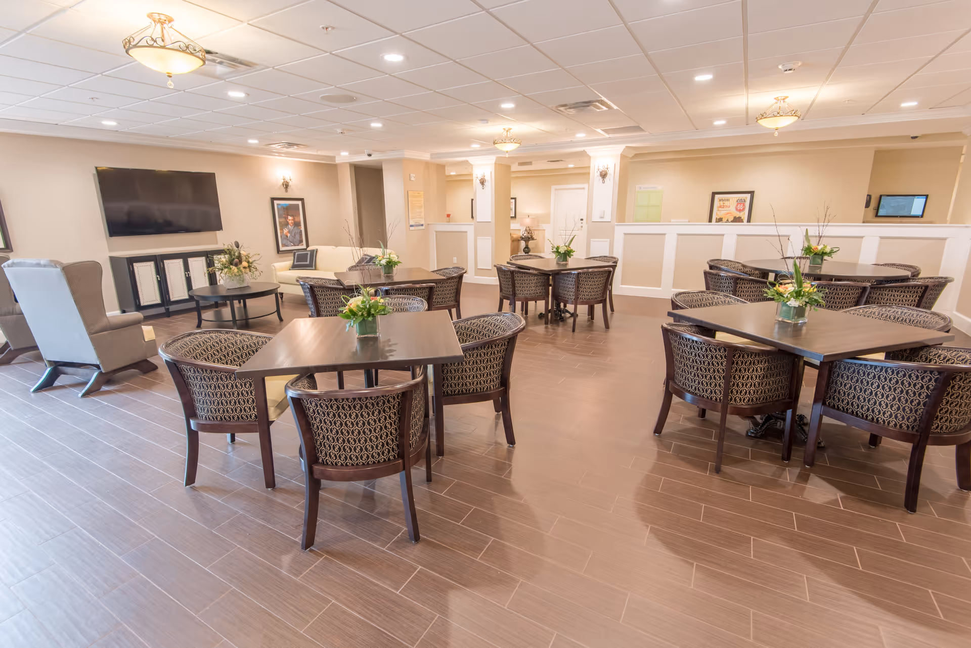 A spacious and well-lit common area with multiple square wooden tables surrounded by patterned cushioned chairs. Each table has a small floral arrangement. The room features a large flat-screen TV mounted on the wall, two armchairs, and light-colored walls with decorative molding. The floor is covered with wood-look tiles, and the ceiling has recessed lighting and decorative light fixtures.