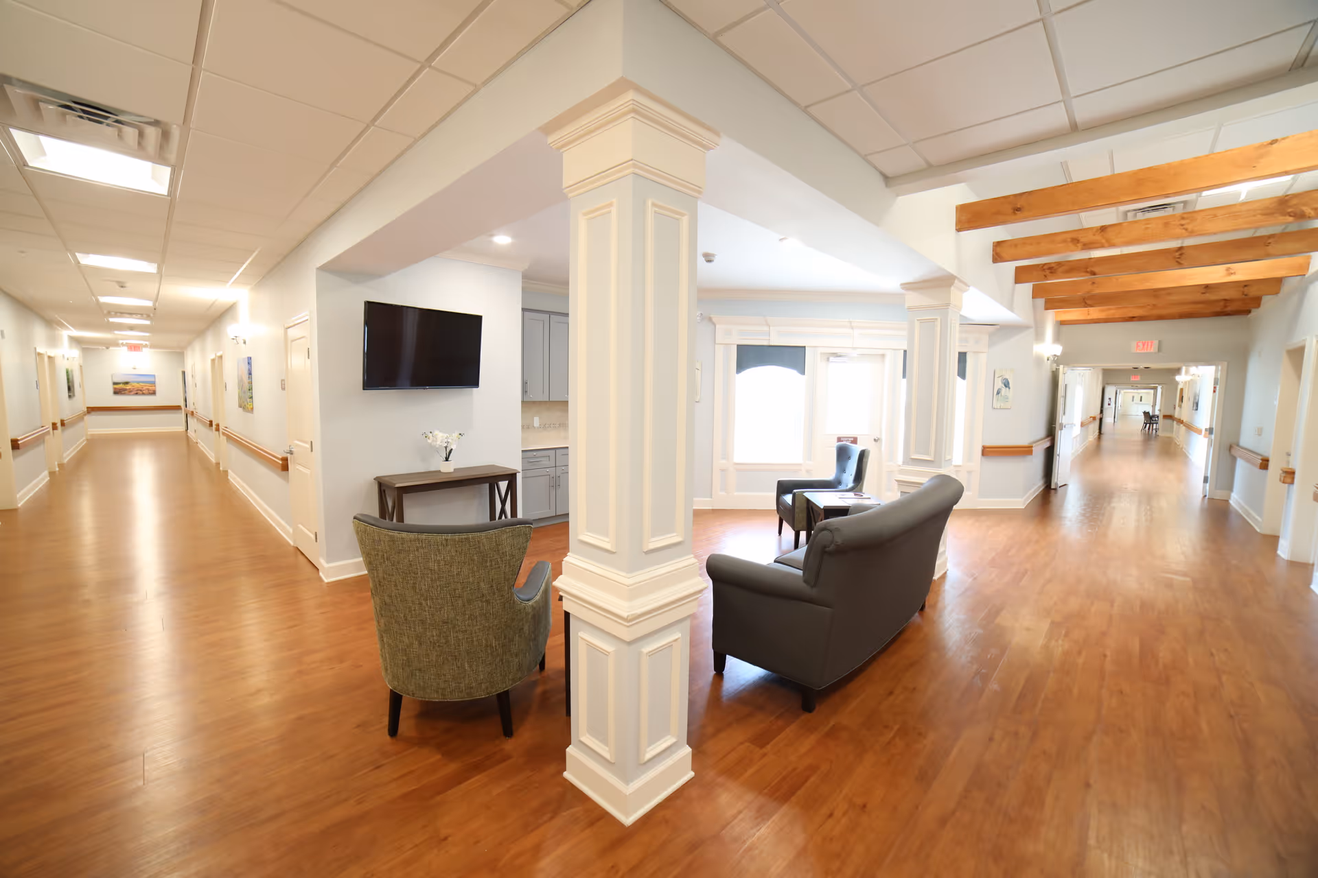 A bright and spacious hallway in a senior living facility with wooden floors and white walls. The hallway features a seating area with two armchairs and a small table near a large window. There is a flat-screen TV mounted on the wall above a small console table with a vase of flowers. The ceiling has recessed lighting and wooden beams on one side. The hallway extends in both directions with doors and handrails along the walls.