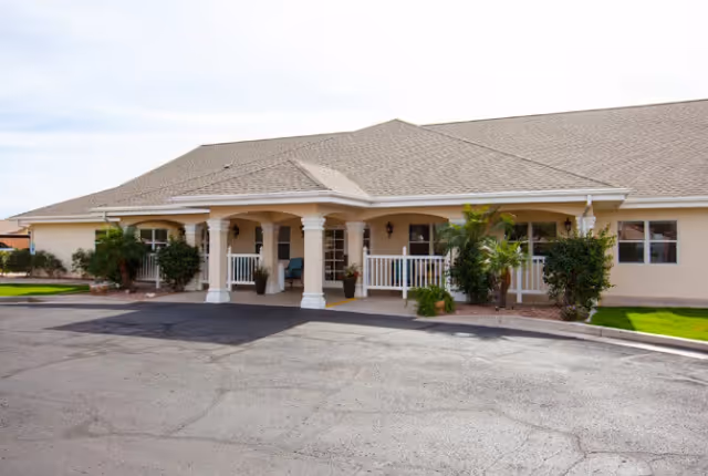 Front exterior view of a single-story senior living facility building with a covered entrance supported by white columns, surrounded by small shrubs and greenery, under a partly cloudy sky.