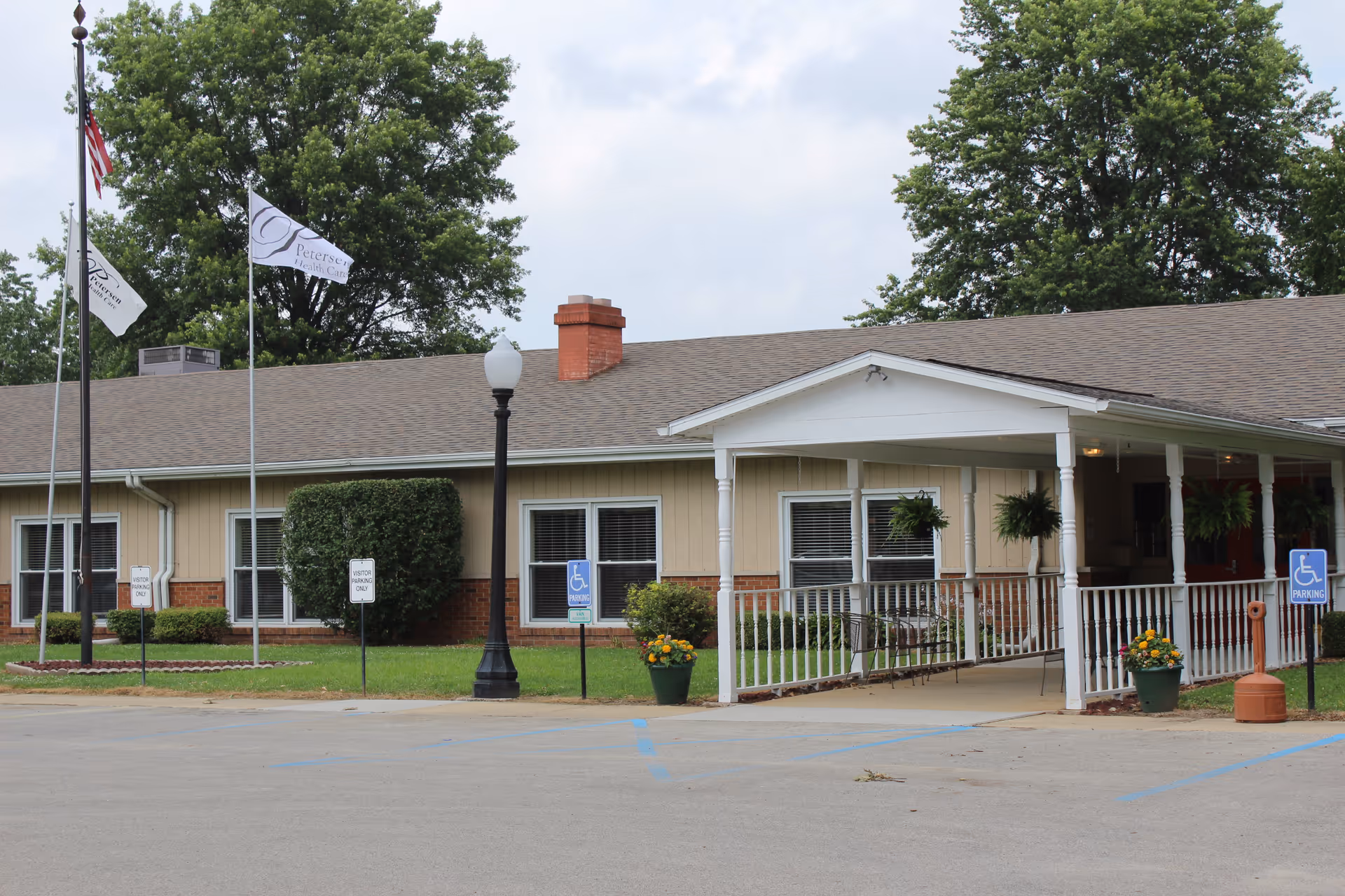 Exterior view of a single-story healthcare facility building with beige siding and a brown roof. There is a covered entrance with a wheelchair accessible ramp, potted plants, and several windows. Three flagpoles with flags are visible on the left side, and there are parking spaces including handicapped spots in front of the building. Trees and a lamppost are also visible in the background.