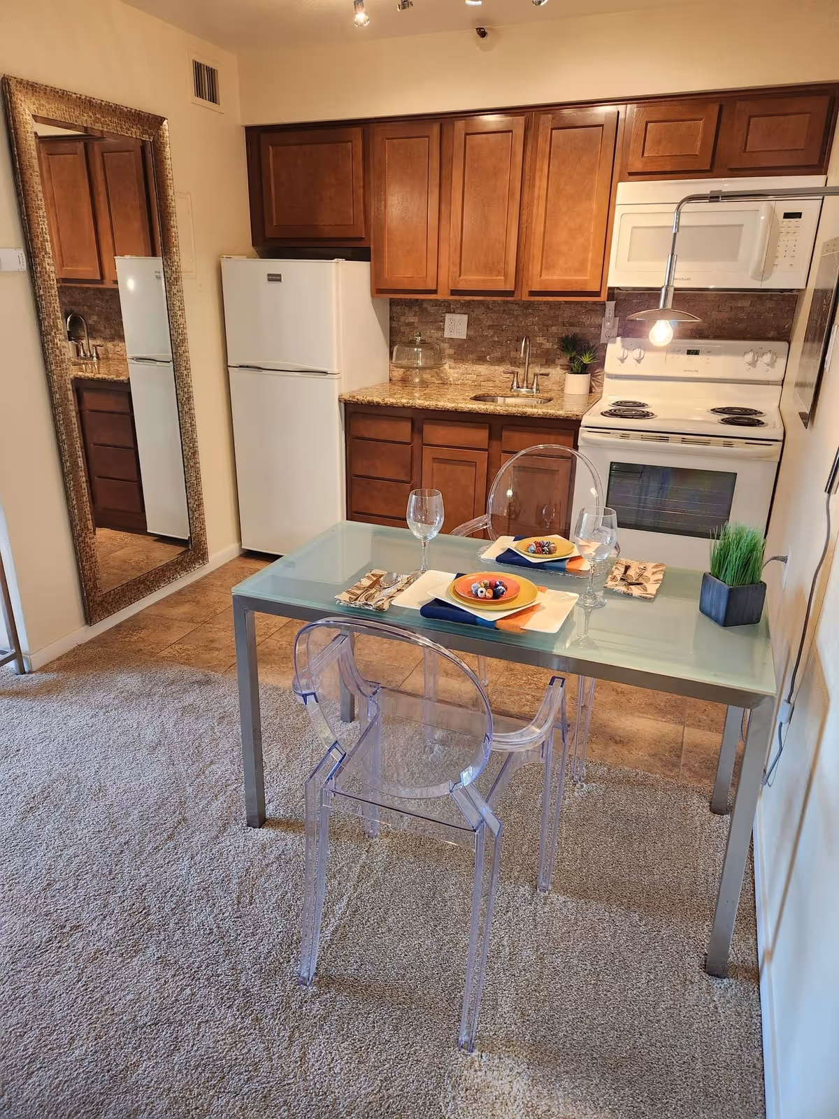 A small kitchen and dining area with wooden cabinets, a white refrigerator, stove, and microwave. A glass dining table is set for two with plates, napkins, and wine glasses. Two transparent chairs are placed at the table. A large decorative mirror is mounted on the wall to the left.