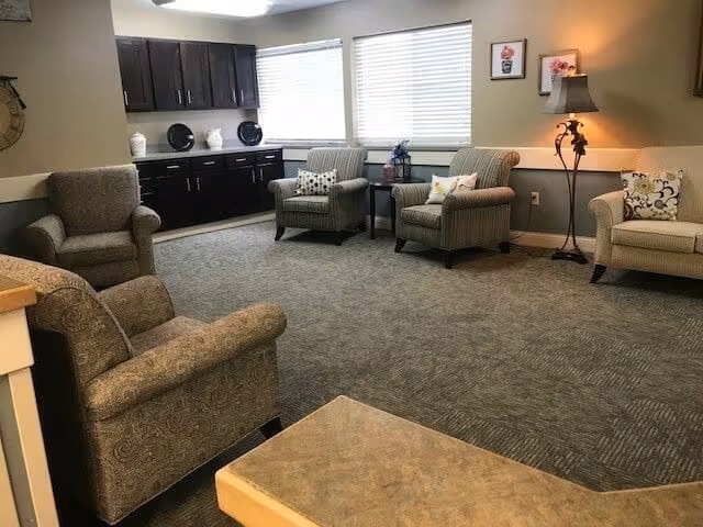 A bright common sitting room with upholstered armchairs arranged on carpet, cabinets along the back wall and windows letting in light.