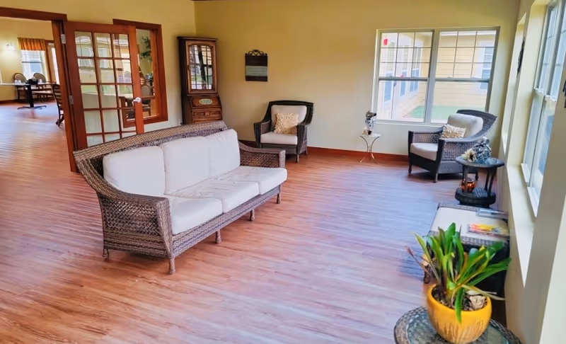 A bright and spacious sitting area with wicker furniture including a sofa and two armchairs with white cushions. The room has wooden flooring, large windows letting in natural light, a small side table with decorative items, and a potted plant in the foreground. Through a glass door, a dining area with tables and chairs is visible.