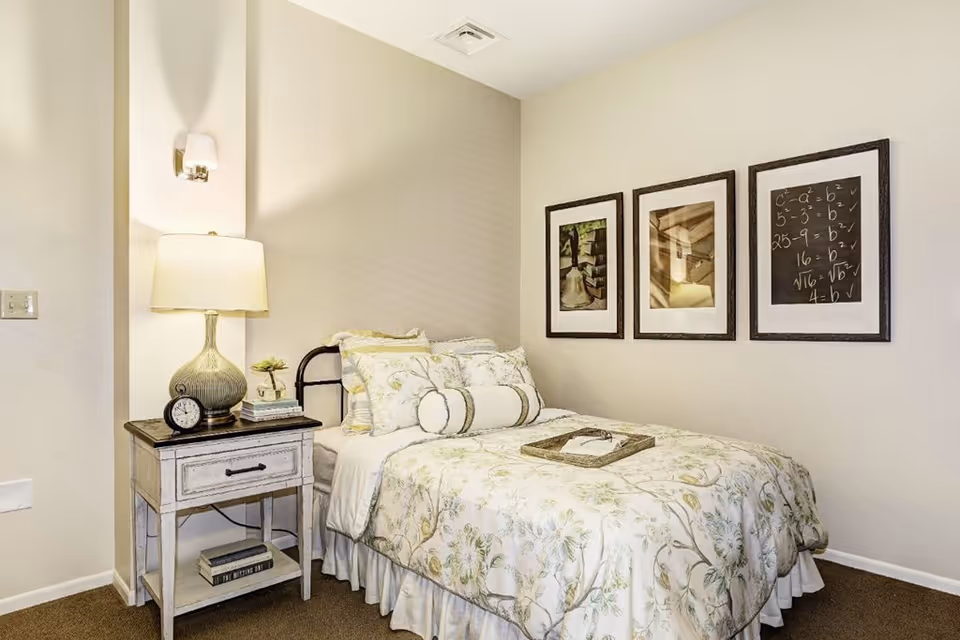 Well-lit single bedroom with a floral bedspread, a nightstand with a lamp and clock, and three framed pictures on the wall.