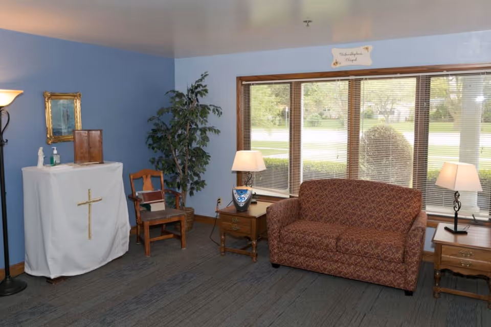 A small chapel room with light blue walls featuring a covered altar with a gold cross, a wooden chair, a potted plant, a patterned sofa, two wooden side tables with lamps, and a large window with blinds showing greenery outside.