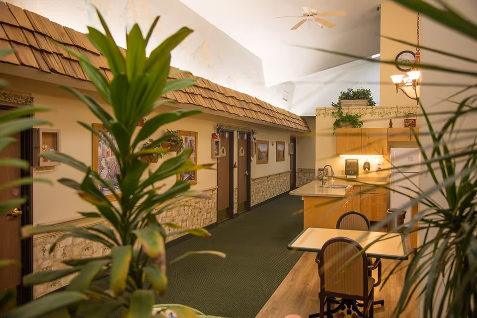 Interior hallway of a senior living facility with a green carpet, wooden doors, and framed pictures on the walls. There is a small kitchenette area with a sink, cabinets, and a refrigerator on the right side, along with a table and chairs. Large green plants partially obscure the view, and the ceiling has a fan and light fixtures.