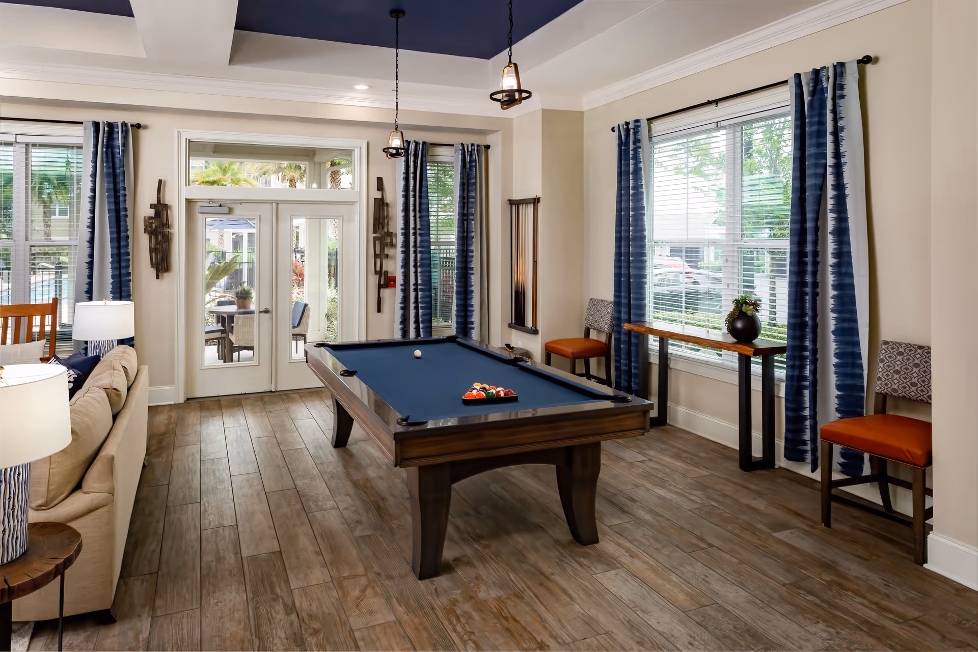 Interior view of a recreational room featuring a pool table with balls arranged for a game. The room has wood-look tile flooring, beige walls, and large windows with blue patterned curtains. There are two chairs and a narrow table against one wall, a beige sofa with a lamp on a side table, and double glass doors leading outside to a patio area with greenery and palm trees visible.