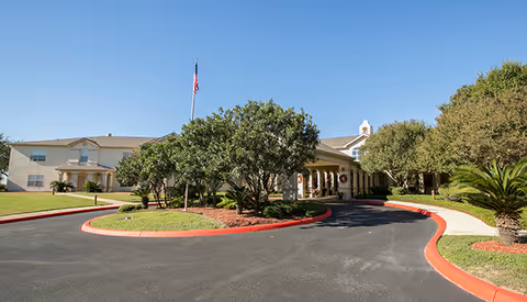 Front exterior view of The William Senior Living facility with a circular driveway, landscaped greenery, trees, and an American flag on a flagpole under a clear blue sky.
