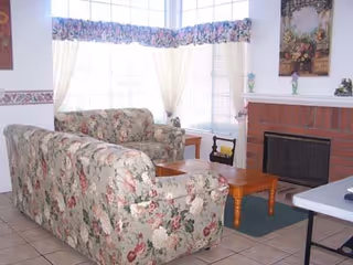 Small living room with matching floral sofas, a wooden coffee table, large bay windows with curtains, and a brick fireplace.