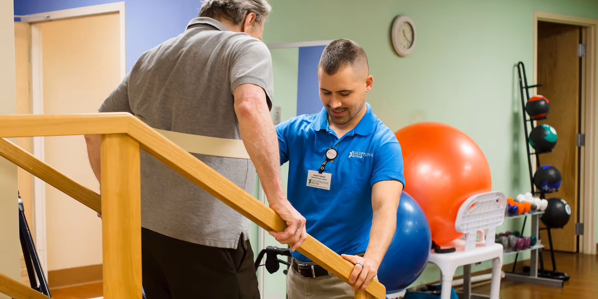 A physical therapist in a blue shirt assists an elderly man using parallel wooden bars for support during a rehabilitation exercise in a therapy room with exercise balls and weights in the background.