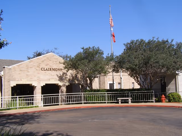 Exterior view of The Clairmont Retirement Community building with a beige brick facade, trees in front, a flagpole with the American and Texas flags, a fire hydrant, and a curved driveway.