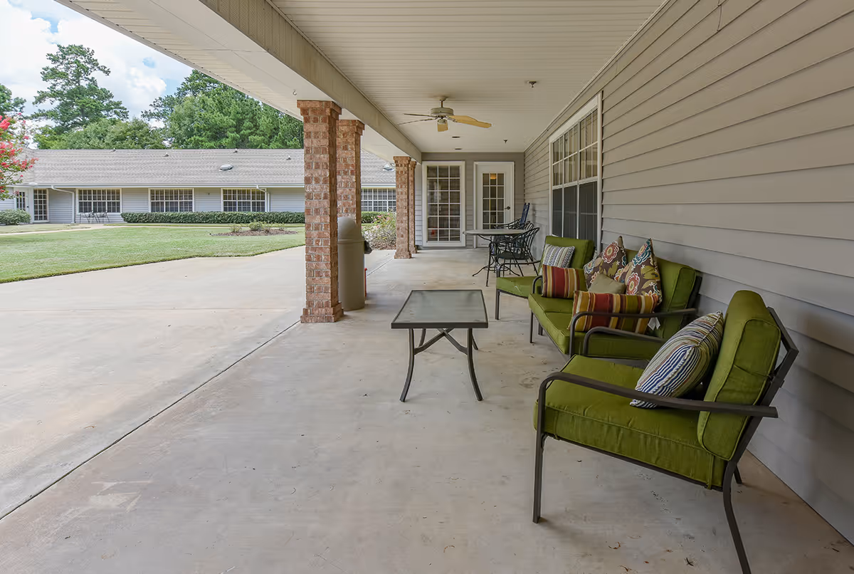 Covered outdoor patio area with green cushioned chairs and a sofa with colorful pillows, a coffee table, and a small dining table with chairs. The patio has brick columns and overlooks a grassy courtyard with another building in the background.