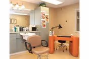 Interior view of a salon area in a senior living facility featuring a salon chair in front of a sink and mirror, a manicure table with a lamp and chair, and cabinets with various supplies.