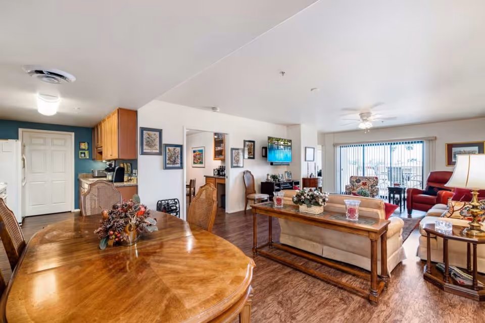 A bright and spacious living and dining area in a senior living facility. The foreground features a polished wooden dining table with chairs and a floral centerpiece. The background shows a cozy living room with a beige sofa, armchairs, a side table with a lamp, a wall-mounted TV, and large sliding glass doors leading to an outdoor patio. The kitchen area with wooden cabinets and appliances is visible to the left.
