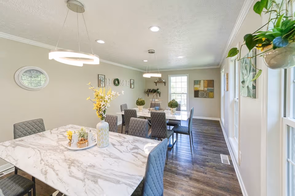 Bright dining room with two marble-patterned tables surrounded by gray quilted chairs. The room features modern circular pendant lights, a wall clock, decorative plants, and artwork on the walls. Large windows on one side let in natural light, and the floor is dark wood.
