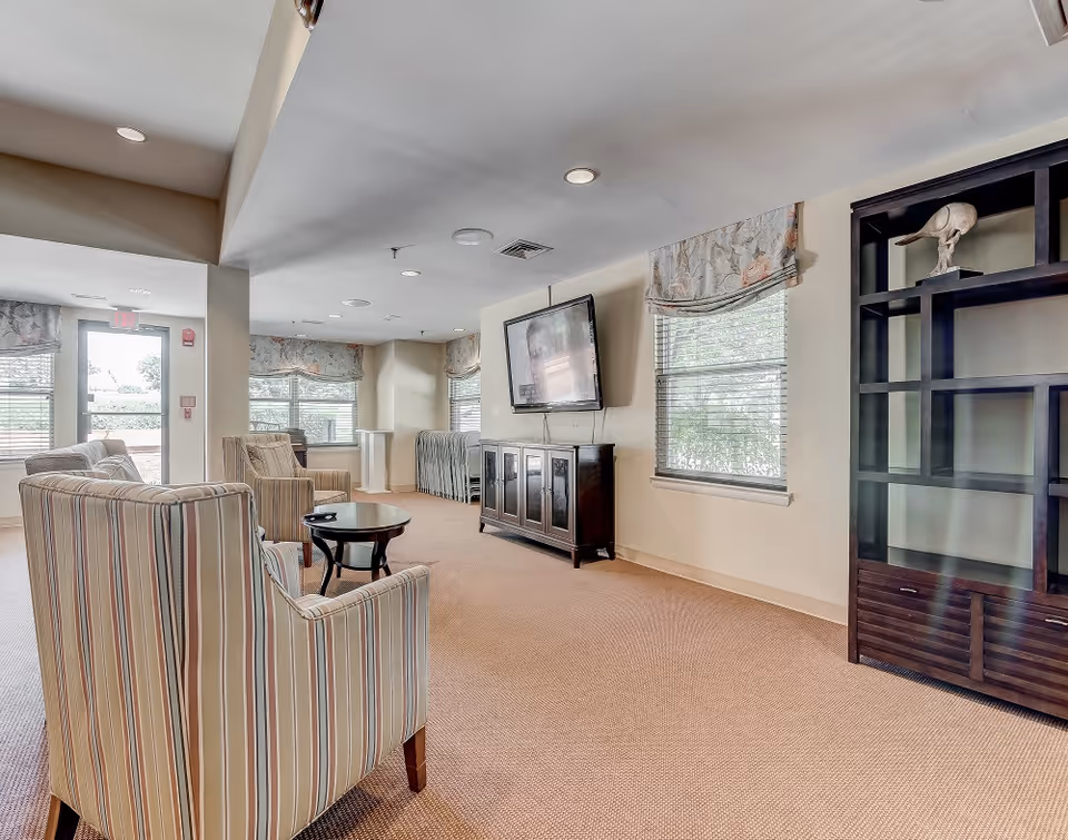 A bright and spacious common area with beige carpet and light-colored walls. The room features striped armchairs arranged around a small round wooden table. A large flat-screen TV is mounted on the wall above a dark wooden cabinet. There are windows with floral valances letting in natural light, and a tall dark wooden shelving unit with decorative items on the right side. The space appears clean and inviting with recessed ceiling lights and an exit door visible in the background.