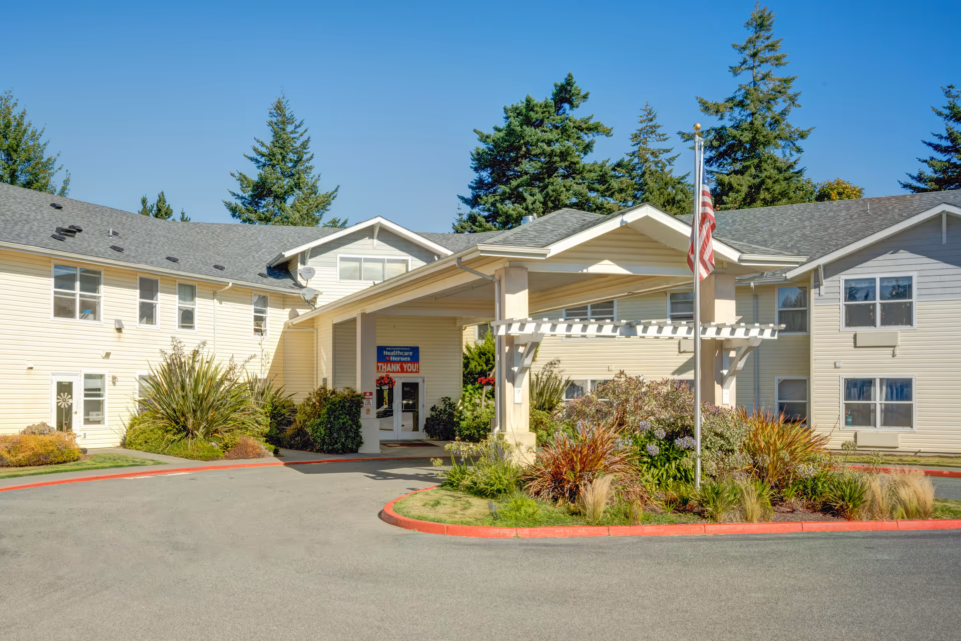 Front entrance of a light-colored senior living building with a covered porte-cochère, landscaped beds, and an American flag.