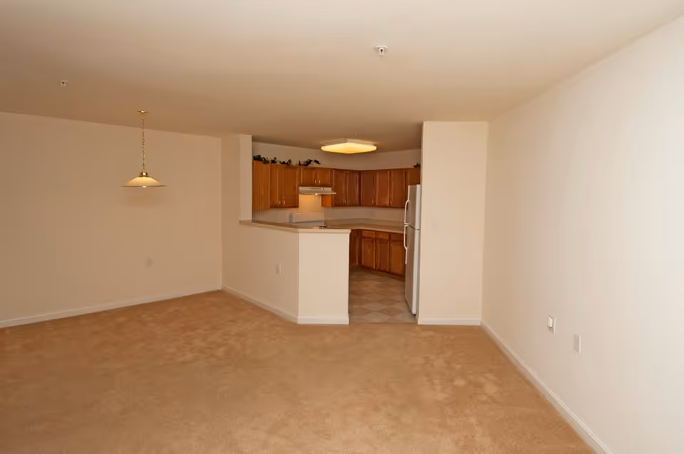 Interior view of an empty apartment showing a beige carpeted living area with white walls, a hanging light fixture, and a kitchen with wooden cabinets, white appliances, and tiled flooring.