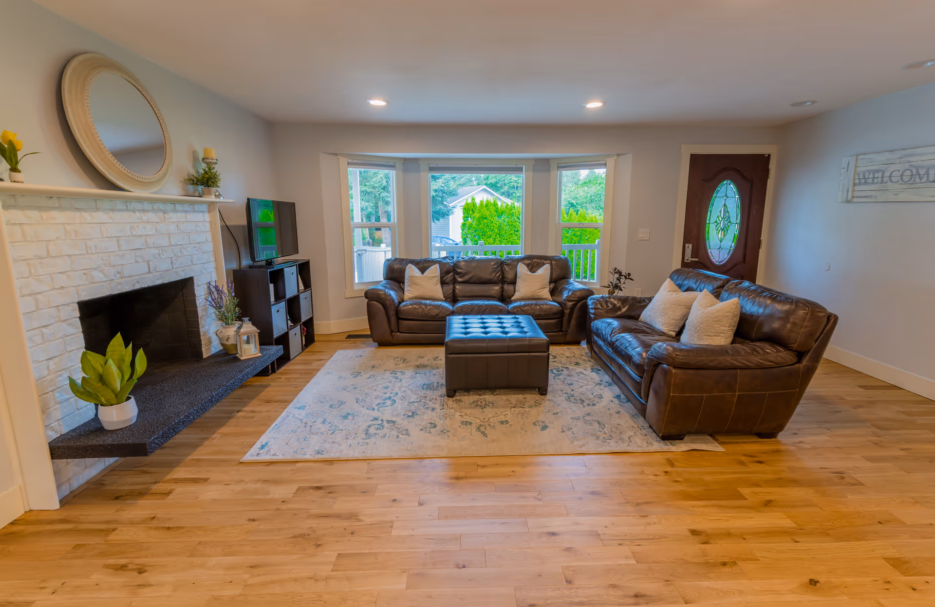 A cozy living room with two brown leather sofas facing each other, a matching ottoman in the center on a patterned area rug, a white brick fireplace with decorative plants and a round mirror above it, a small TV on a black stand, and a wooden front door with an oval glass window. Large windows behind the sofas let in natural light and show greenery outside.