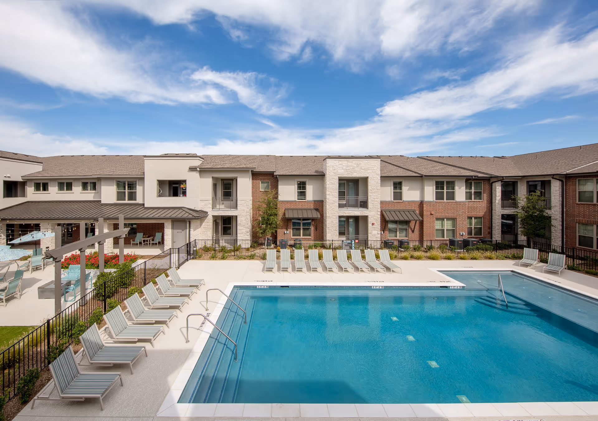 Outdoor swimming pool area at The Luxe at Rowlett with multiple lounge chairs arranged around the pool. The pool is surrounded by a fence and there is a two-story building with balconies and windows in the background under a partly cloudy blue sky.