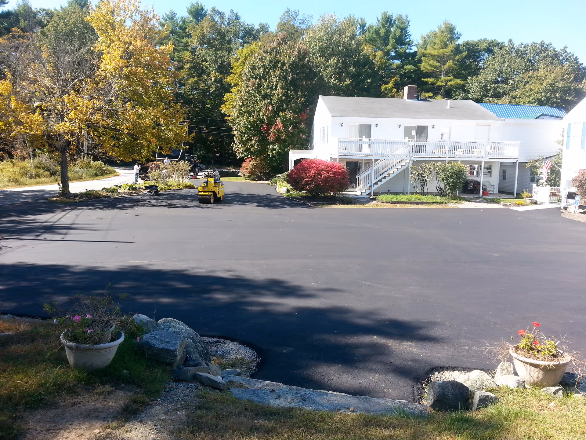 A newly paved parking area in front of a white two-story building with a balcony and stairs leading to the upper level. The scene includes trees with autumn foliage, potted plants on either side of the paved area, and a small yellow road roller vehicle on the asphalt. The background shows a wooded area under a clear blue sky.