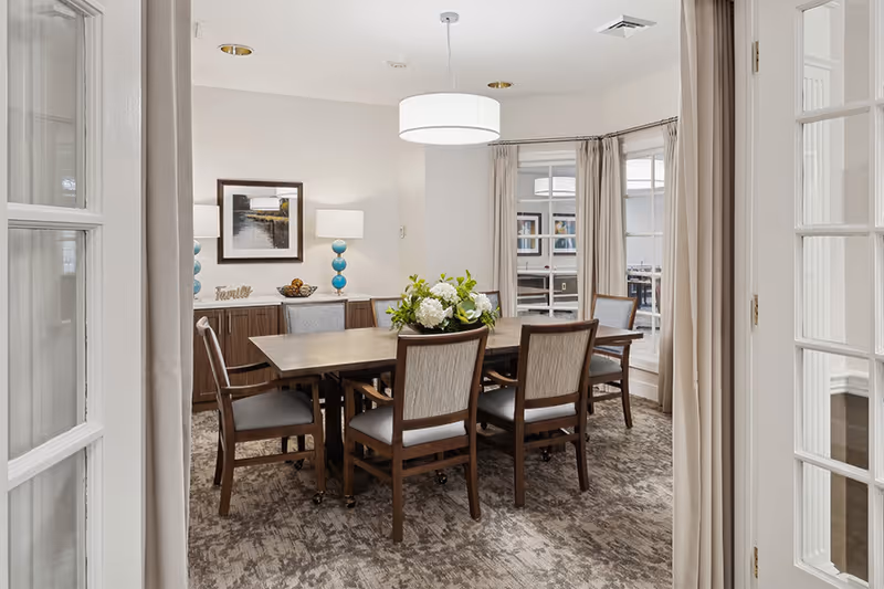 A well-lit dining room with a rectangular wooden table surrounded by six cushioned chairs. A floral centerpiece decorates the table. The room features large windows with beige curtains, a modern ceiling light fixture, a sideboard with two blue lamps, a framed picture on the wall, and a carpeted floor.