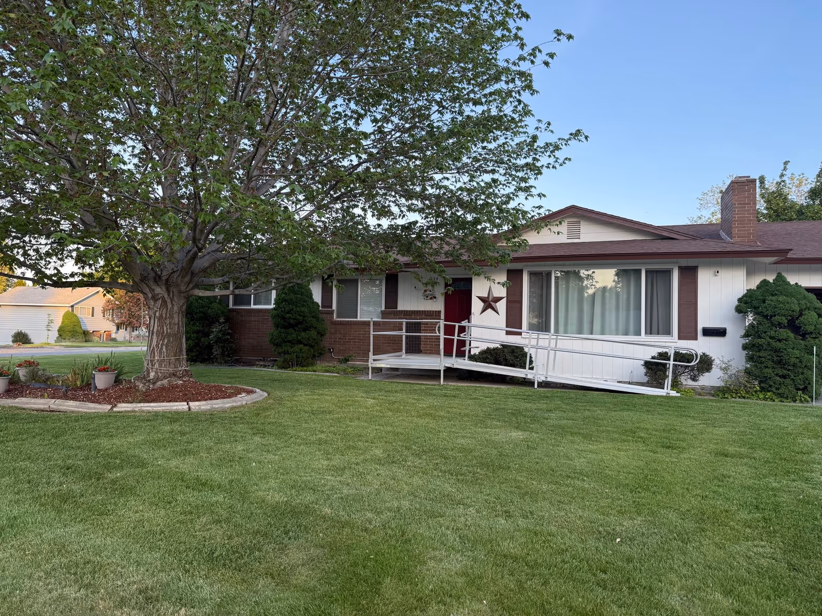 Front exterior of a single-story house with a wheelchair ramp, a large tree, and a manicured lawn.