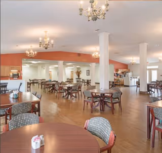 Spacious dining room with multiple round tables and chairs, chandeliers, and a serving area in the background.