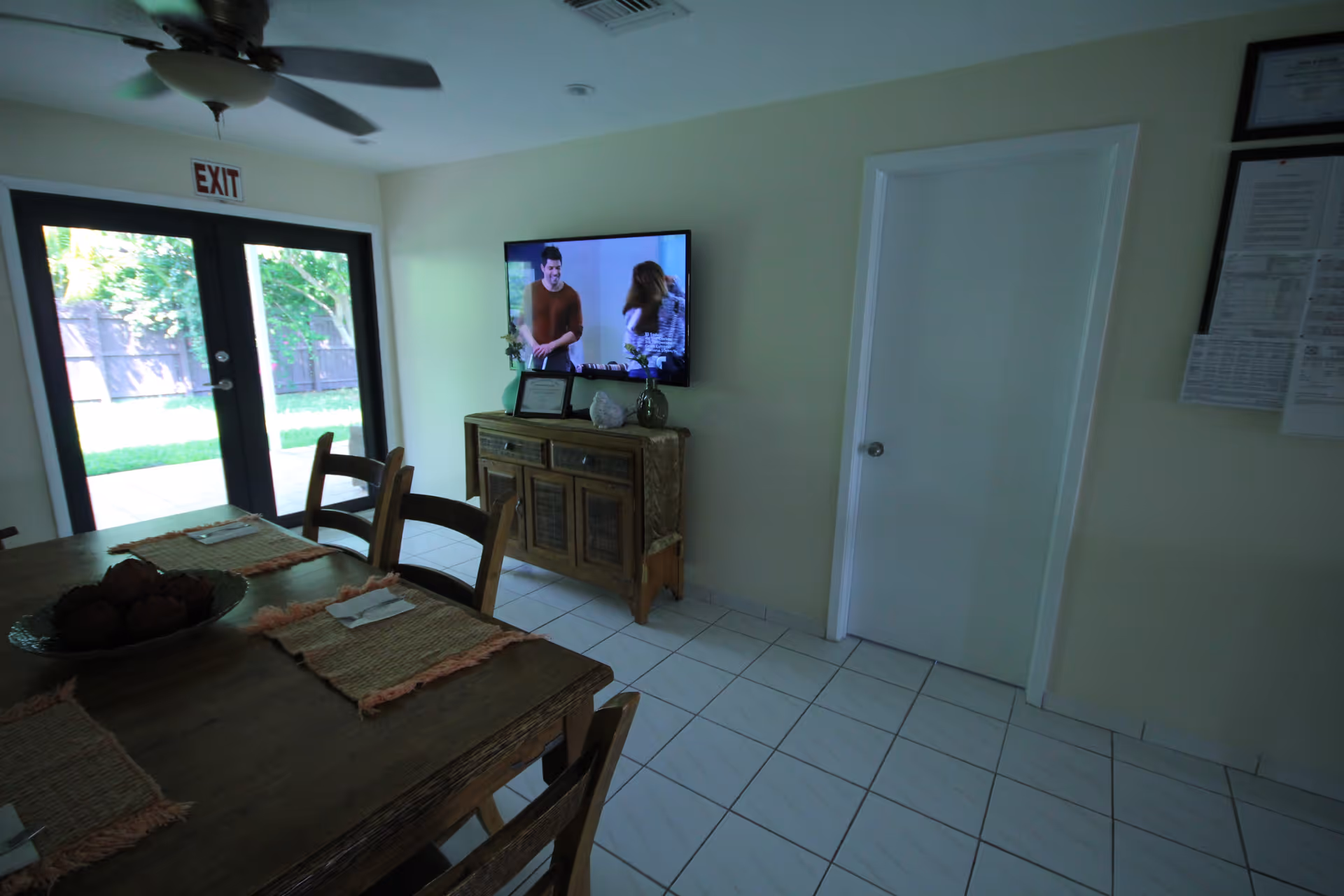 A dining area with a wooden table and chairs, placemats on the table, a ceiling fan above, a TV mounted on the wall showing two people, a wooden cabinet below the TV, a door to the right, and glass double doors leading outside with an exit sign above them.