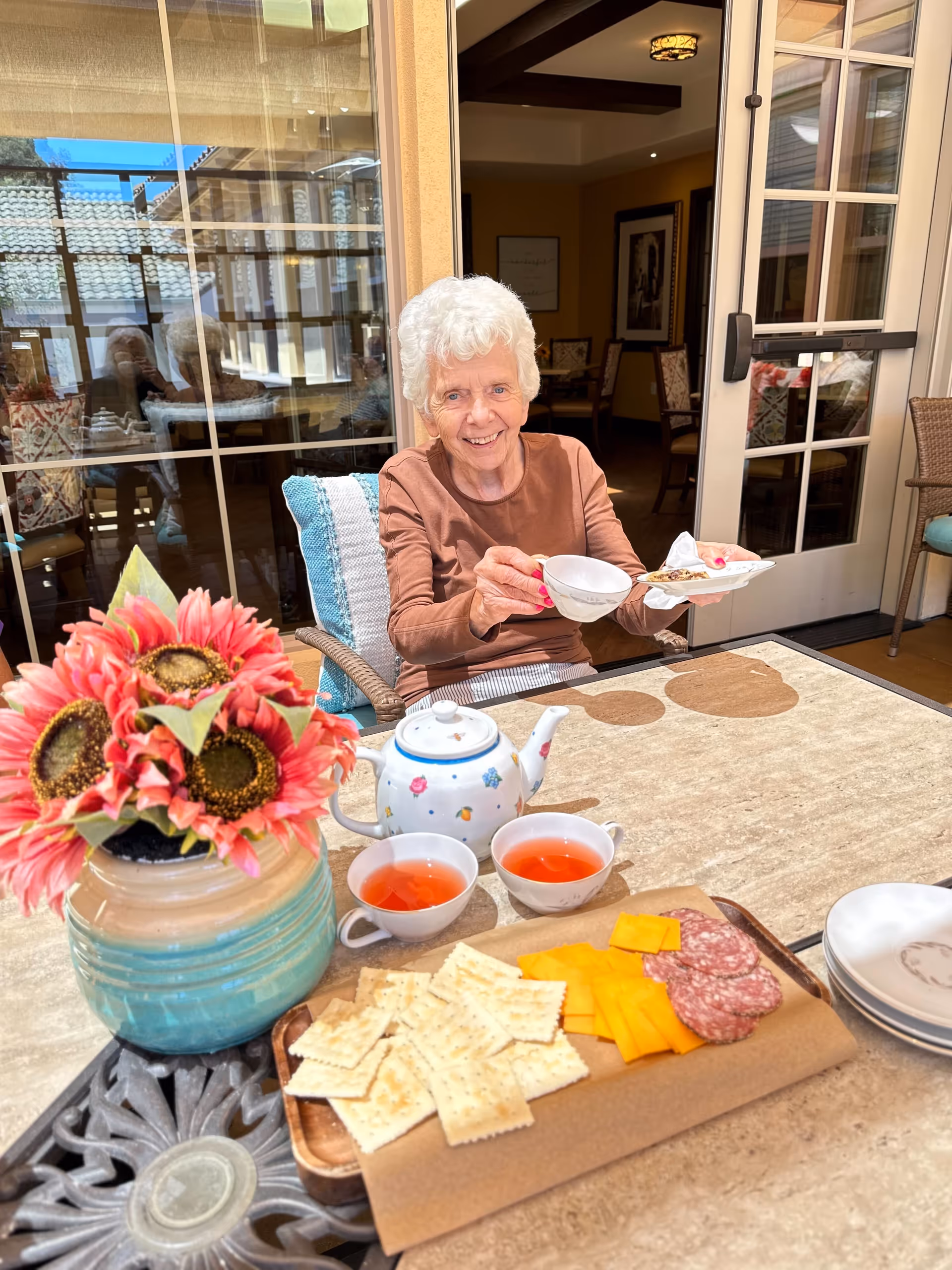 An elderly woman with white hair sitting at a table outdoors, smiling and holding a teacup and a small plate with a snack. On the table in front of her are two cups of tea, a teapot, a wooden tray with crackers, cheese, and slices of salami, and a vase with bright pink sunflowers. Behind her is a glass door leading to an indoor dining area.